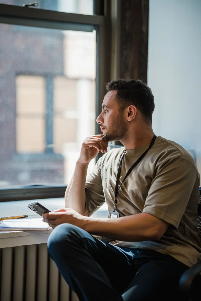 Homme dans un bureau est assis en tailleur près d'une fenêtre, plongé dans ses pensées tout en tenant un téléphone portable